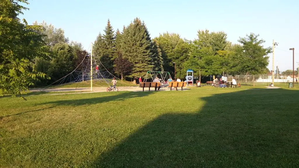 Parc verdoyant avec aire de jeux, structures d’escalade, balançoires et familles profitant du soleil en plein air